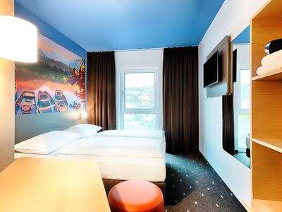 Hell eingerichtetes Hotelzimmer mit Doppelbett, buntem Teppich, Fenster mit Blick und moderner Wandbeleuchtung.