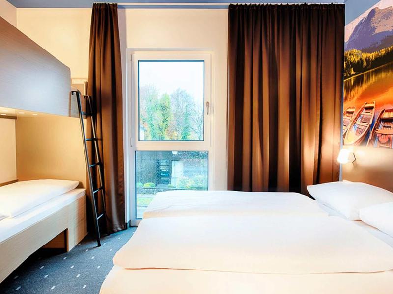 Modernes Hotelzimmer mit Doppelbett, Sitzbank, dunklen Vorhängen und Landschaftsbild an der Wand.