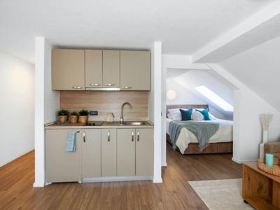 Kleine Küche mit beige Schränken, Spüle und Holzboden, angrenzend an Schlafzimmer unter Dachschräge.
