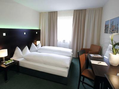 Habitación de hotel con dos camas individuales, escritorio, sillas y ventana con cortinas beige.