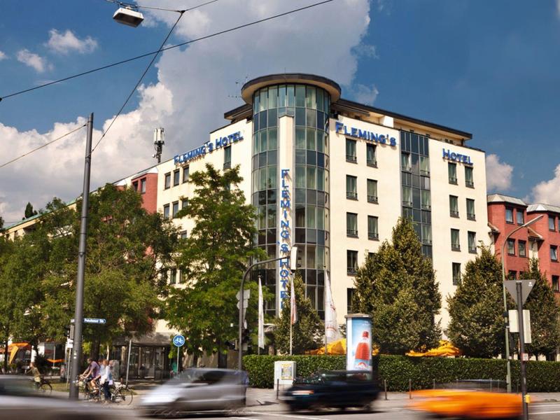 Hotel urbano moderno con ascensor de vidrio en varios pisos, rodeado de árboles y tráfico callejero.