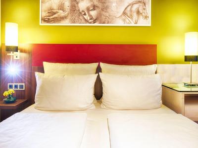 Moderne hotelkamer met twee eenpersoonsbedden, rood hoofdeinde en gele muren, verlicht door twee lampen.