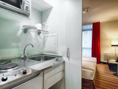 Kleine hotelkeuken met gootsteen, kookplaat en magnetron, naast slaapkamer met rode gordijn.