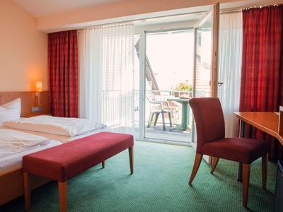 Jasny pokój hotelowy z dwoma łóżkami, czerwoną ławką i dostępem do balkonu.