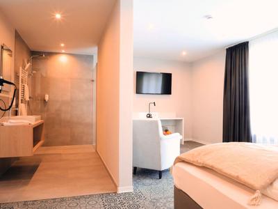 Nowoczesny pokój hotelowy z otwartą łazienką, fotelem, telewizorem i dużymi oknami z zasłonami.