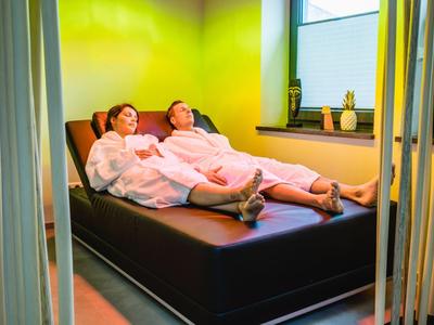 Dwie osoby w szlafrokach relaksują się na czarnym leżaku przy zielono oświetlonej ścianie.