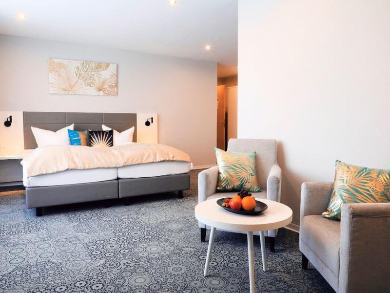 Jasno urządzone pokoje hotelowe z podwójnym łóżkiem, fotelami i małym stolikiem z owocami.