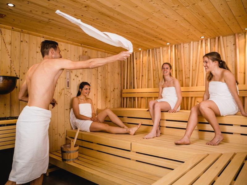 Cztery osoby relaksują się w drewnianej saunie, trzy siedzą, jedna wachluje ręcznikiem.