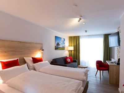 Modernes Hotelzimmer mit zwei Einzelbetten, Sofa, Schreibtisch und großem Fenster mit grünen Vorhängen.