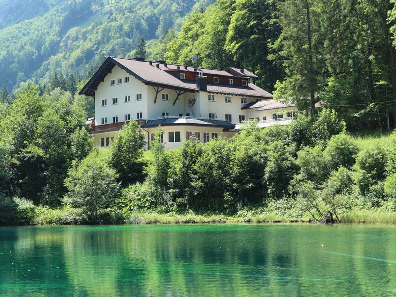 Hotel in een groene bosomgeving bij een helder meer met bergen op de achtergrond.