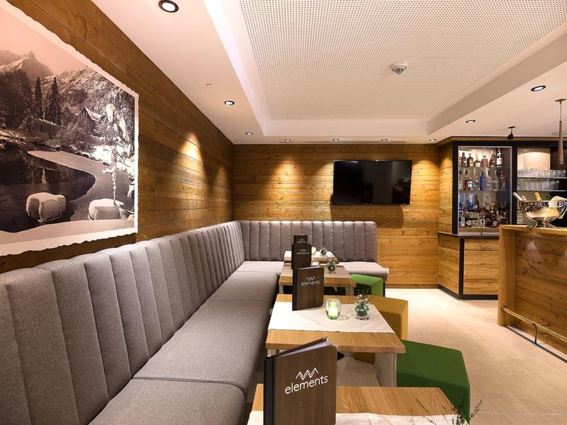 Gezellig hotel-loungedeel met grijze gestoffeerde bank, houten tafels en moderne verlichting.