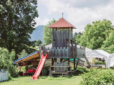 Een speelplaats voor kinderen met een houten toren en een rode glijbaan omgeven door bomen.