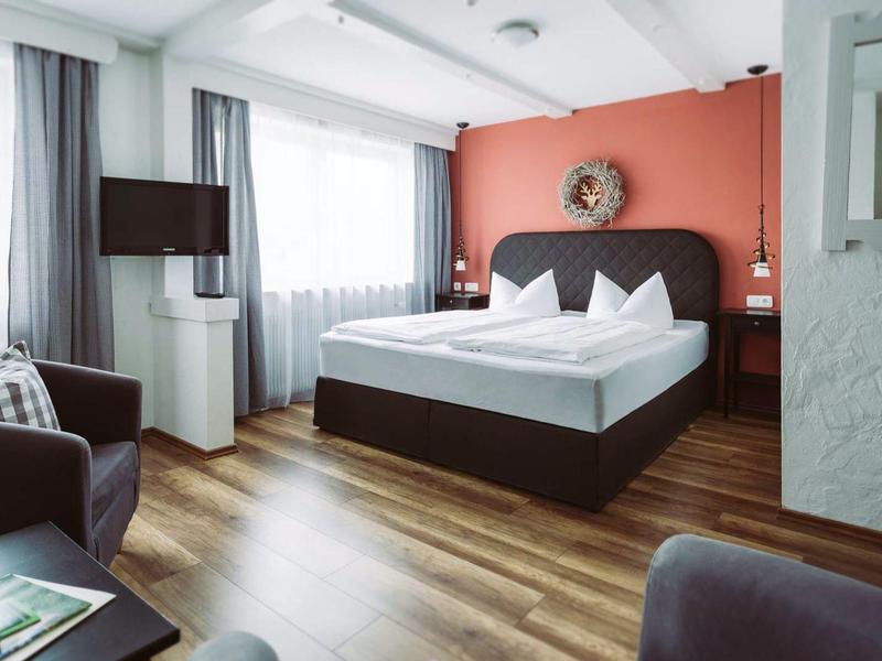 Lichte hotelkamer met tweepersoonsbed, houten vloer en warme rode accentmuur.