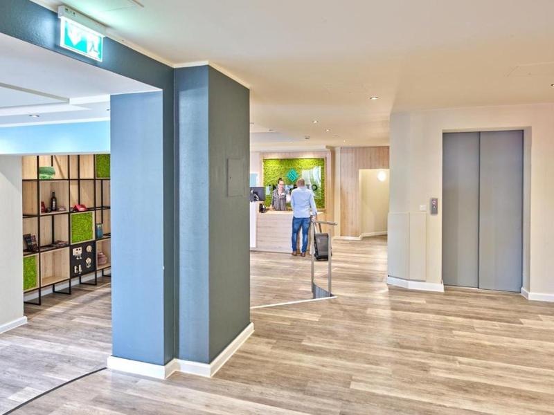 Modern hotel corridor with elevators, wooden floor, and illuminated emergency exit sign.