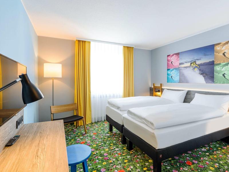 Camera d'hotel luminosa con due letti singoli, tende gialle, moquette decorata e scrivania con TV.