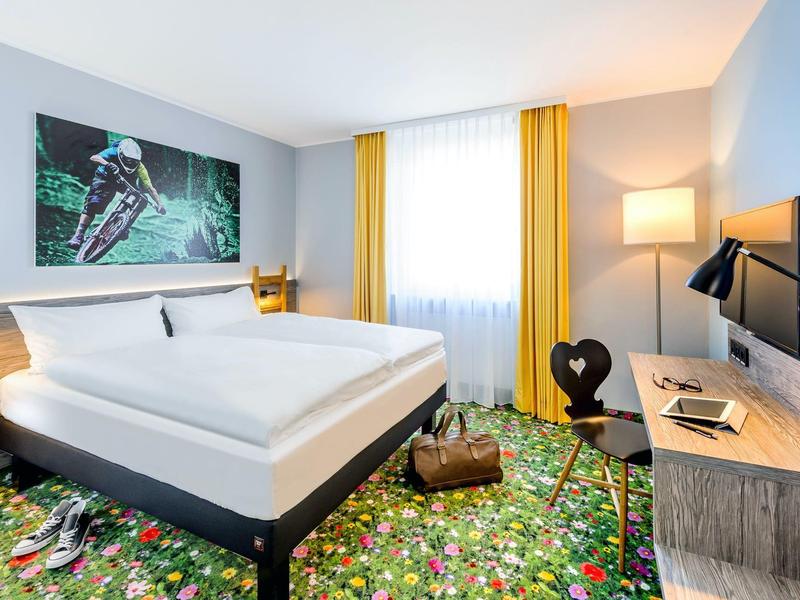 Camera d'hotel luminosa con letto matrimoniale, moquette floreale colorata, tende gialle e TV a parete.