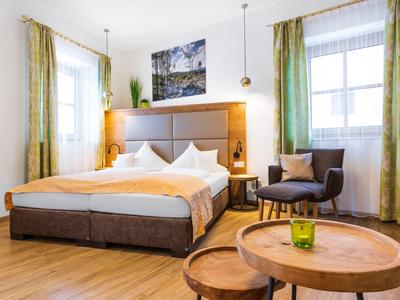 Světle zařízený hotelový pokoj s manželskou postelí, křesly a dřevěným stolem před okny se zelenými závěsy.