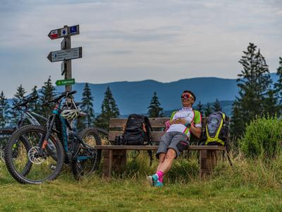 Osoba odpočívá na lavičce vedle dvou kol s horskou krajinou v pozadí.