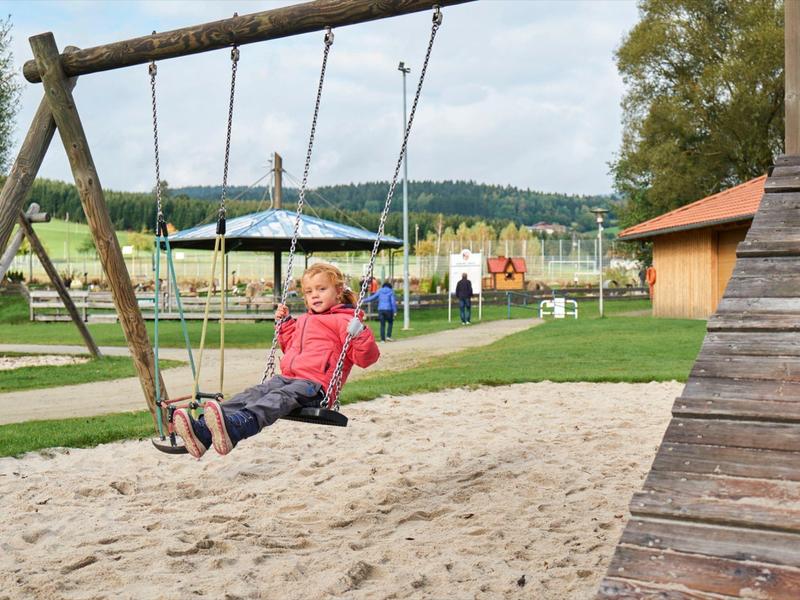 Bambino dondola su un'area sabbiosa del parco giochi con paesaggio rurale sullo sfondo.