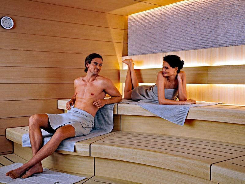 Due persone rilassate in una sauna di legno con asciugamani, che si sfidano a braccio di ferro.