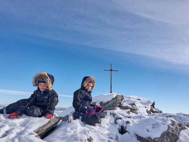 Due bambini seduti nella neve accanto a una croce di vetta sotto il cielo azzurro.