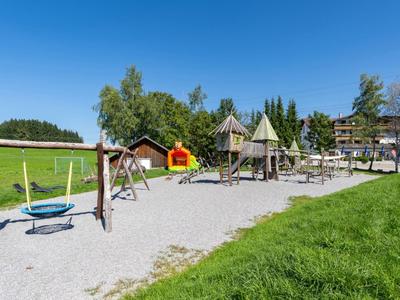 Aire de jeux avec balançoires et structures d'escalade sur gravier à côté d'une pelouse verte sous un ciel clair.