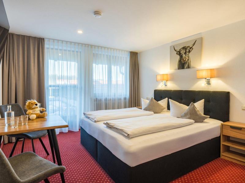 Chambre d'hôtel lumineuse avec lit double, table, chaise et grandes fenêtres avec rideaux.