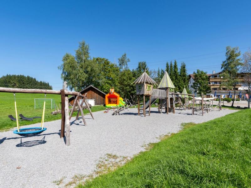 Aire de jeux avec balançoires et structures d'escalade sur gravier à côté d'une pelouse verte sous un ciel clair.