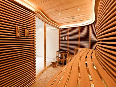 Moderne Sauna mit geschwungenen Holzbänken und Wandpaneelen, warmes Licht, heller Boden.