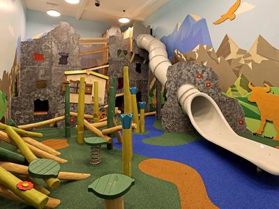 Bunter Indoorspielplatz mit Klettergerüst, Rutsche und Wald- und Bergmotiven an den Wänden.