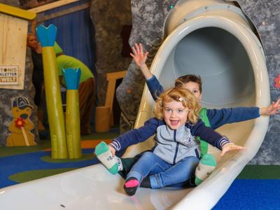 Zwei Kinder rutschen lachend aus einer weißen Rutsche in einer bunten Indoor-Spielhalle.