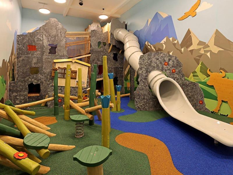 Bunter Indoorspielplatz mit Klettergerüst, Rutsche und Wald- und Bergmotiven an den Wänden.