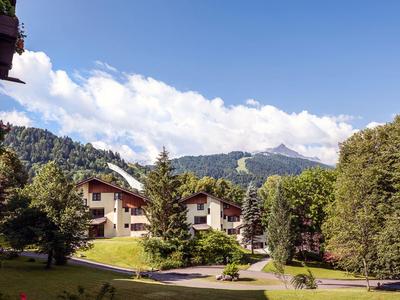 Bergdorf mit weißen Häusern, grünen Bäumen und Wiesen unter blauem Himmel mit Wolken.