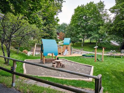 Speelplaats voor kinderen met speelhuisjes en klimtoestellen in een groen park naast een houten hek.