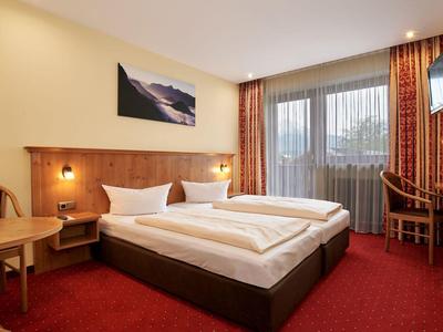 Lichte hotelkamer met tweepersoonsbed, bureau, tv en balkon met gordijnen.