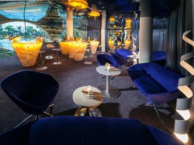 Moderní hotelový bar s modrými křesly, kulatými stoly a výrazným osvětlením.