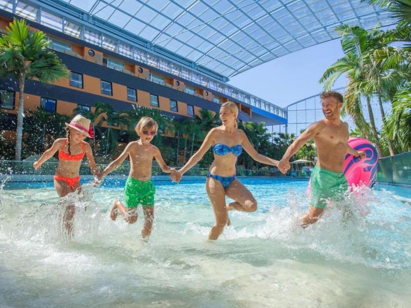 Rodina se dvěma dětmi běží a směje se v krytém bazénu se skleněnou střechou.