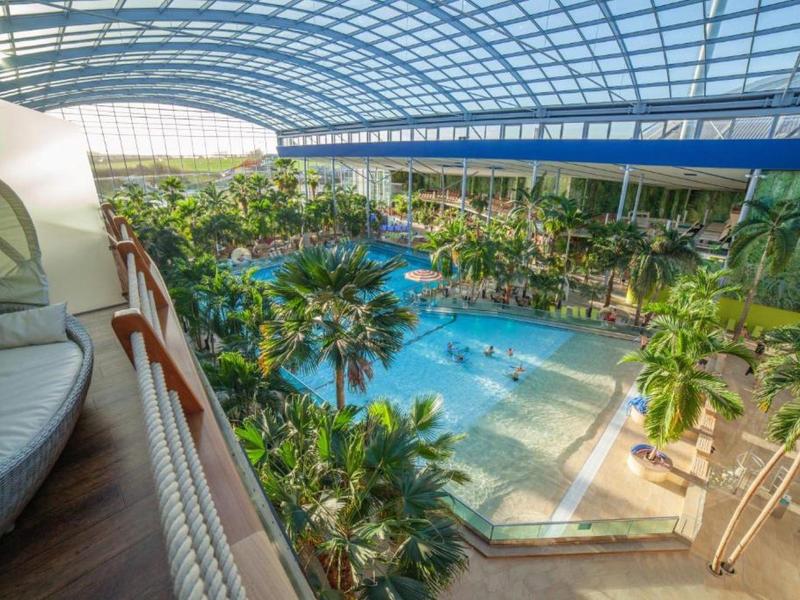 Prostorná vnitřní bazénová oblast s tropickými rostlinami a odpočinkovou zónou pod skleněnou střechou.
