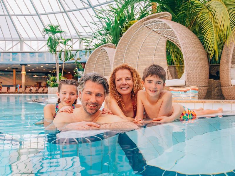 Rodina se dvěma dětmi si užívá bazén v tropickém krytém vodním parku s palmami a lehátky.