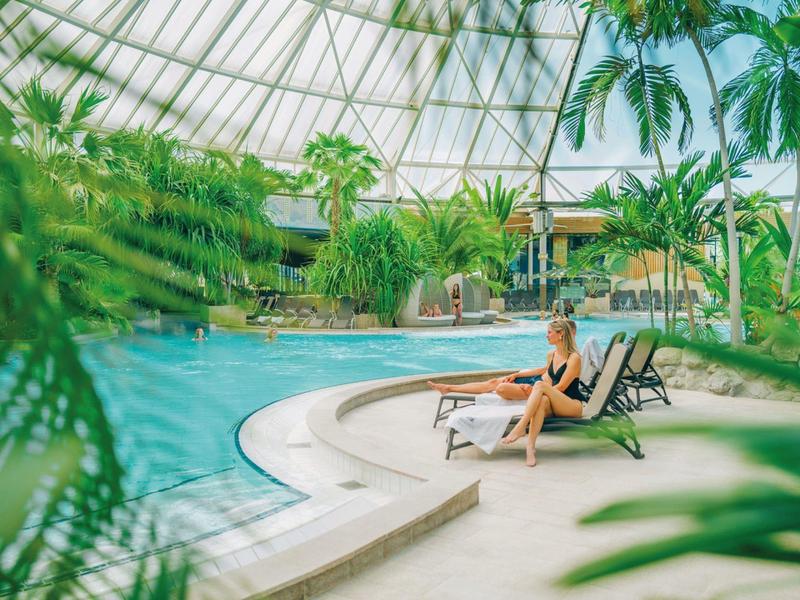 Žena odpočívá na lehátku u bazénu v tropickém hotelovém prostředí pod skleněnou střechou.
