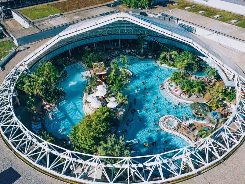 Kruhový venkovní bazén s tobogány, vodními atrakcemi a bujnou zelení, obklopený budovou.