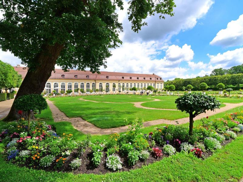 Großer Garten mit Blumenbeeten und altem Baum vor einem langen Gebäude bei sonnigem Himmel.