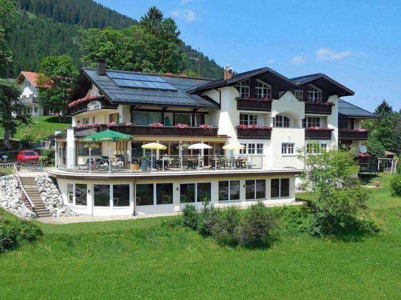 Grand hôtel avec terrasse ensoleillée dans un paysage montagneux vert sous un ciel bleu.