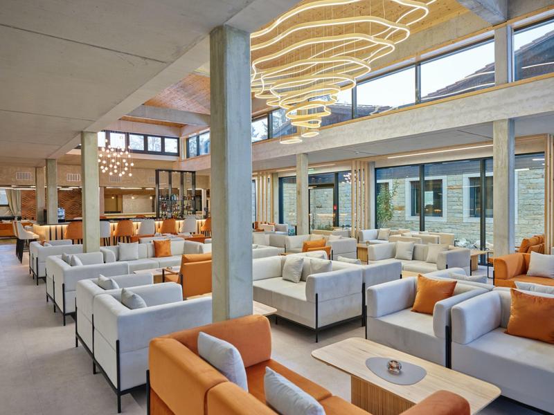 Nowoczesny hotelowy salon z białymi i pomarańczowymi meblami oraz dużymi oknami.