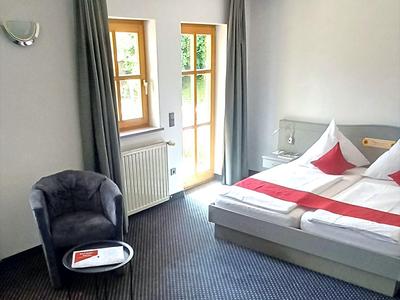 Camera d'hotel luminosa con letto matrimoniale, poltrona grigia e accesso al balcone tramite ampie finestre.