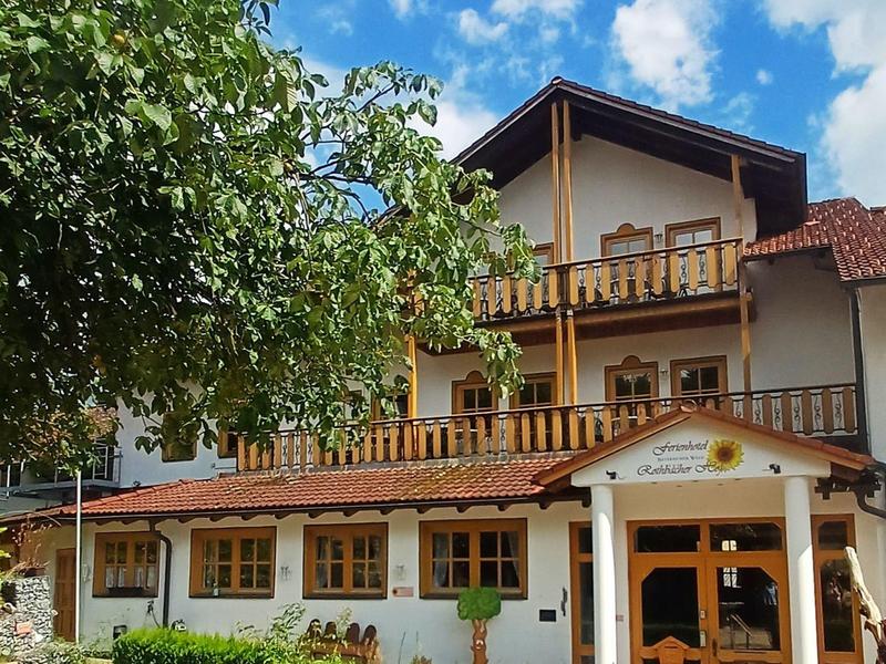 Wielopiętrowy hotel z balkonami, otoczony zielonymi krzewami i drzewem pod niebieskim niebem.