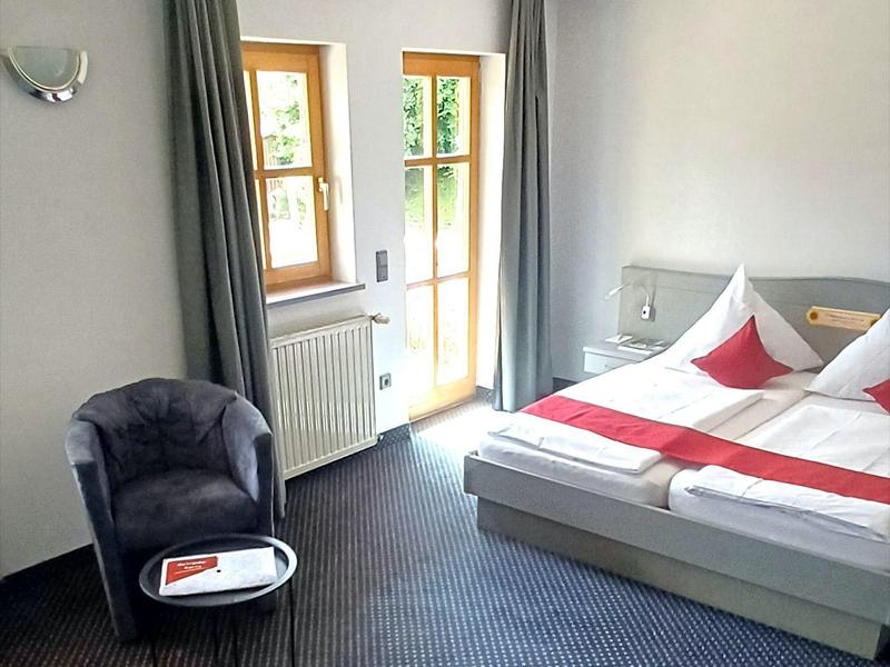 Jasno urządzone hotelowe pokoje z podwójnym łóżkiem, szarym fotelem i oknem na taras.