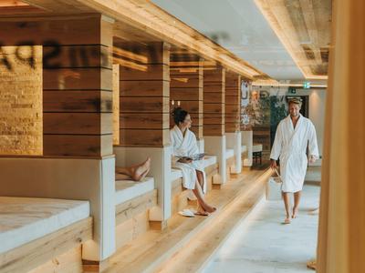 Zwei Personen in weißen Bademänteln entspannen in einem modernen Spa mit Holzwänden und Liegen.