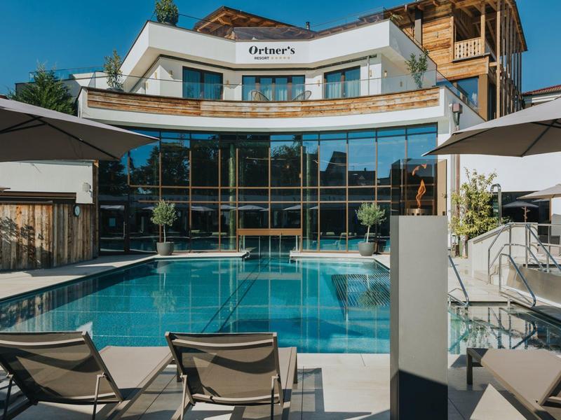 Moderner Hotelpool mit Liegestühlen und Sonnenschirmen vor einem mehrstöckigen Gebäude bei klarem Himmel.