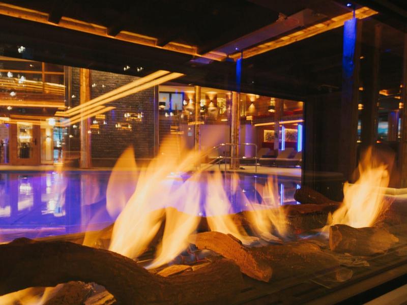 Moderne Feuerstelle in einer Hotel-Lobby mit Glaswänden und beleuchtetem Pool im Hintergrund.